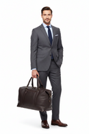 A man in a gray suit, white shirt, and navy tie stands against a white background, holding a dark brown barbour leather duffel bag.