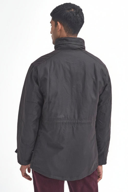 A man viewed from the back wearing a barbour sapper wax rustic jacket with a high collar and a drawstring waist, paired with burgundy pants, against a plain white background.