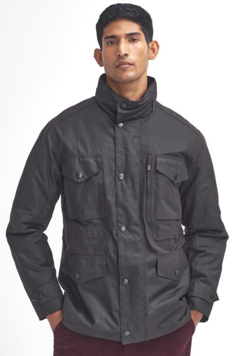 A man wearing a barbour sapper wax rustic jacket with a high stand-up collar and multiple front pockets, paired with burgundy corduroy pants, against a plain white background.