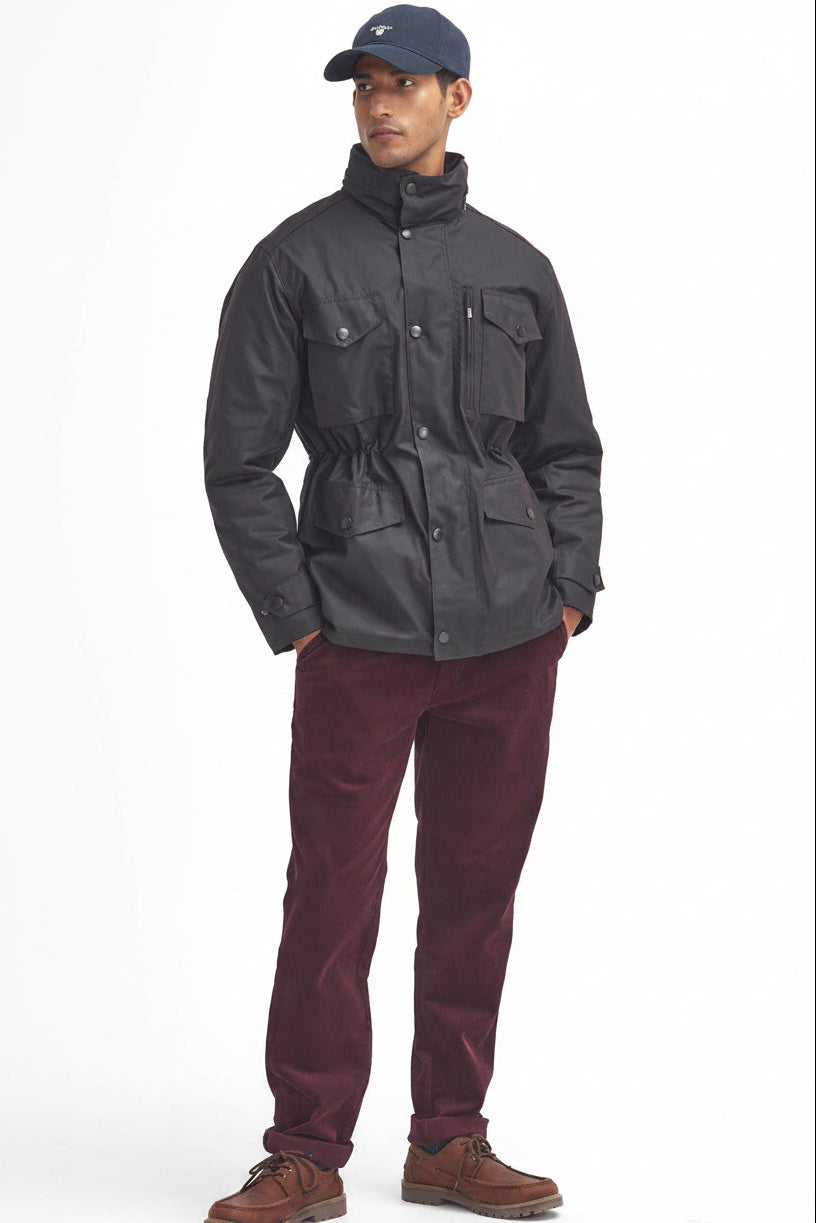 A man modeling a barbour sapper wax rustic jacket with a high collar, paired with burgundy corduroy trousers and brown boots, against a plain white background.